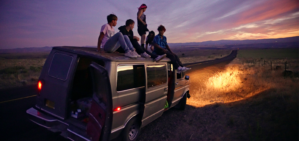 Cinco jovens estão sentados em cima do teto de uma van, que tem os faróis acesos. Eles estão em uma estrada erma, no fim da tarde.