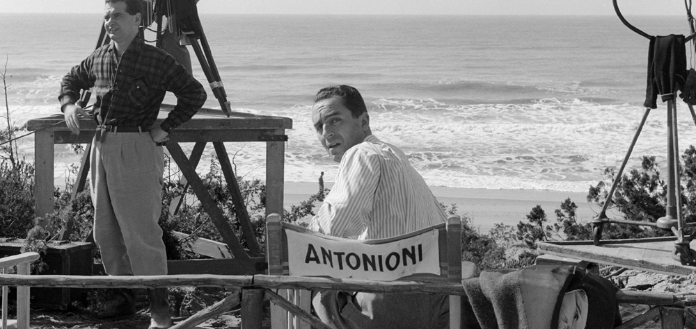 Michelangelo Antonioni está sentando em frente ao mar, em uma cadeira de diretor que tem seu nome. Ele está olhando para trás, em direção à câmera.